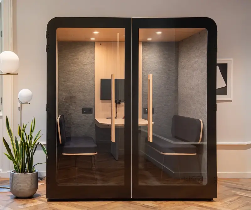 Cabine acoustique moderne en verre avec une table, un écran mural, deux sièges rembourrés gris, plante verte et lampadaires dans un bureau.