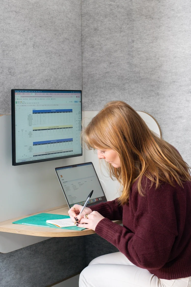 Collaboratrice prenant des notes dans une cabine acoustique, avec écran affichant un tableau de reporting
