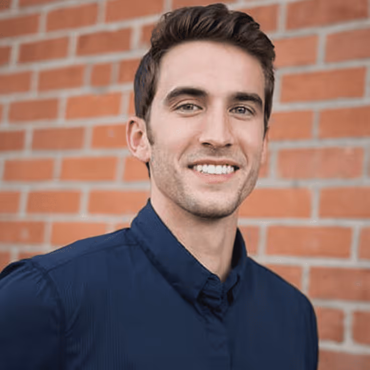 Young man with short brown hair wearing a navy blue shirt smiling in front of a brick wall.