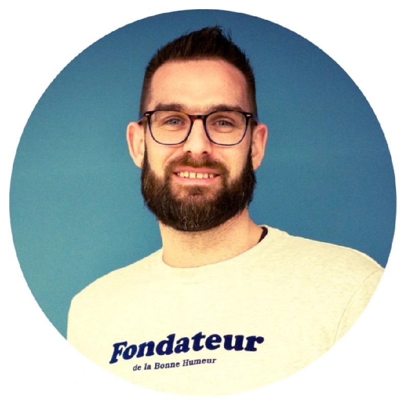 Smiling man with glasses and beard wearing a white shirt with 'Fondateur de la Bonne Humeur' text on a blue background.