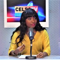Woman with long black hair wearing a yellow sweater speaking into a microphone in a radio studio.