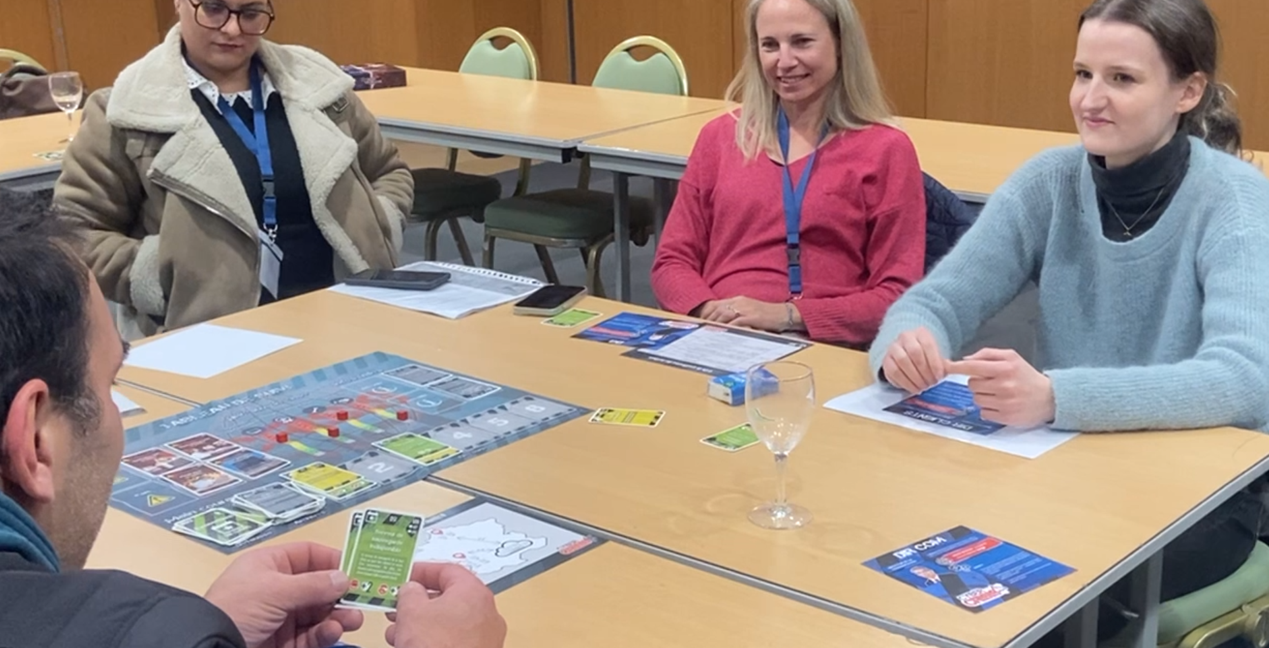 Four adults seated around a table playing a board game with cards and game pieces.