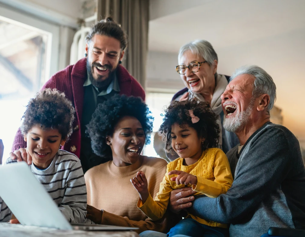 Une famille multigénérationnelle souriante autour d'un ordinateur portable dans un salon lumineux.