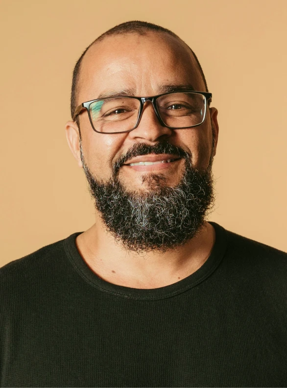 Homme souriant avec une barbe grise, portant des lunettes et un t-shirt noir, devant un fond beige.