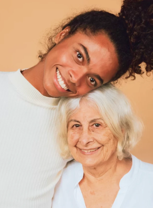 Jeune femme souriante appuyant la tête sur celle d'une femme âgée souriante devant un fond beige.