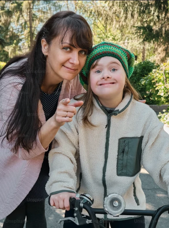 Une femme souriante en veste rose pointe vers l'appareil photo à côté d'une jeune fille avec trisomie 21 portant un chapeau vert et un sweat beige, tenant un vélo.