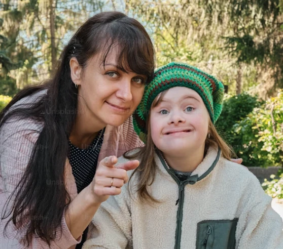 Une femme aux cheveux longs bruns sourit et pointe vers la caméra tandis qu'une jeune fille portant un bonnet vert sourit à ses côtés en extérieur.