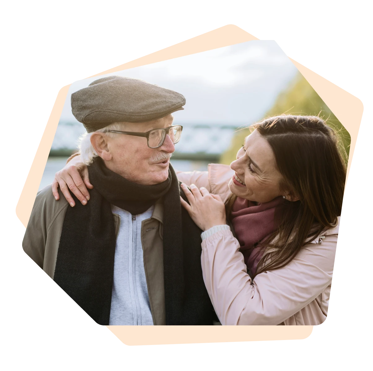 Un homme âgé portant une casquette et une écharpe noire sourit à une femme souriante qui a les mains posées sur ses épaules.