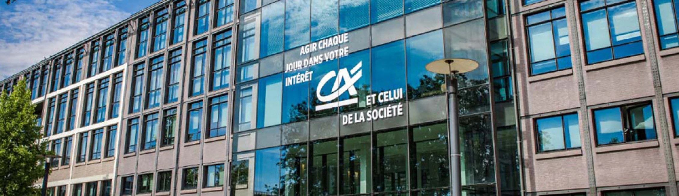 Façade moderne en verre d'un bâtiment de bureau avec le logo Crédit Agricole et le slogan « Agir chaque jour dans votre intérêt et celui de la société ».
