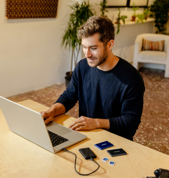 Un homme souriant travaille sur un ordinateur portable à une table en bois avec des disques durs externes et des cartes mémoire devant lui.