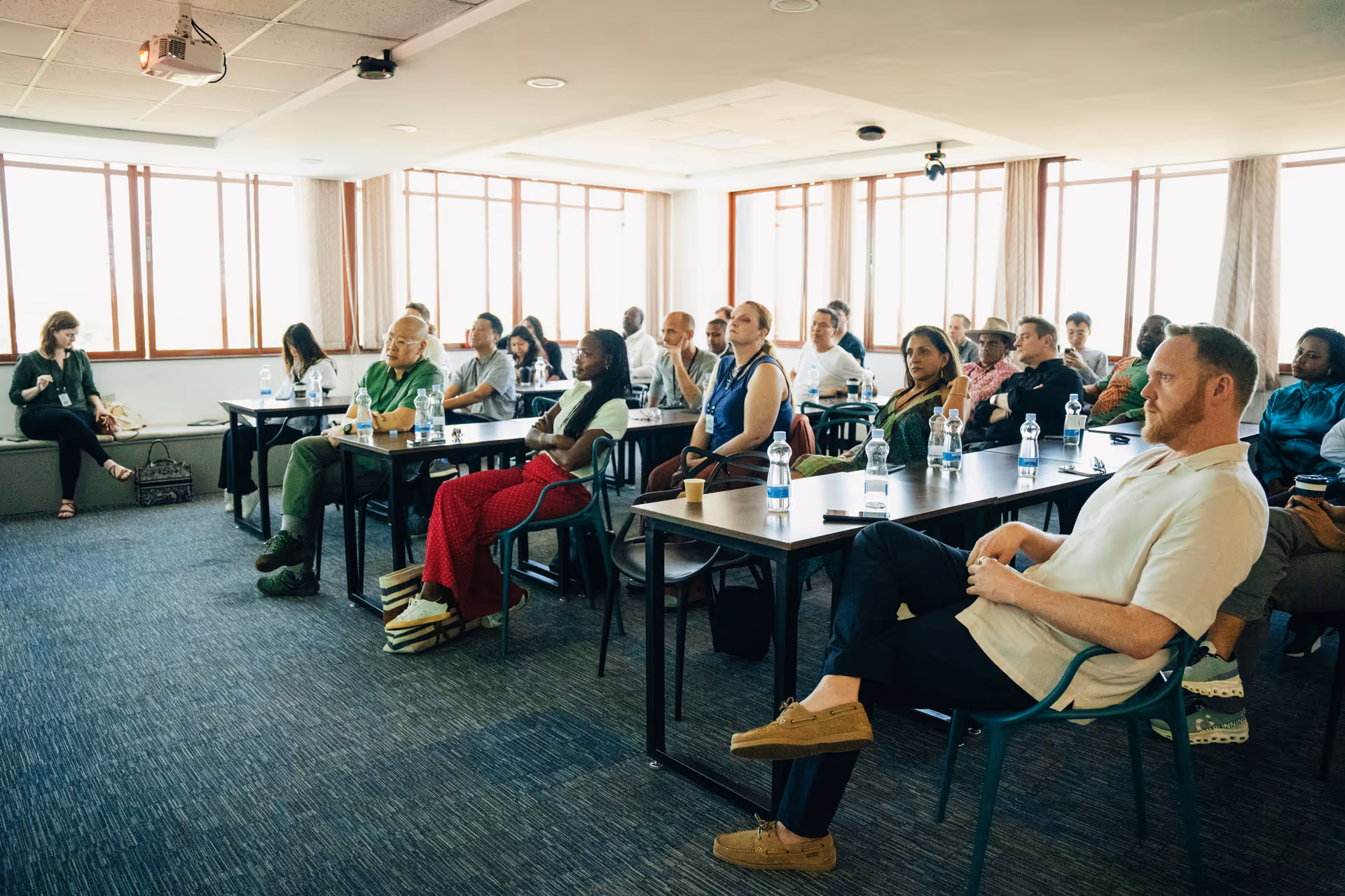 Diverse adults seated and attentively listening in a bright conference room with large windows.