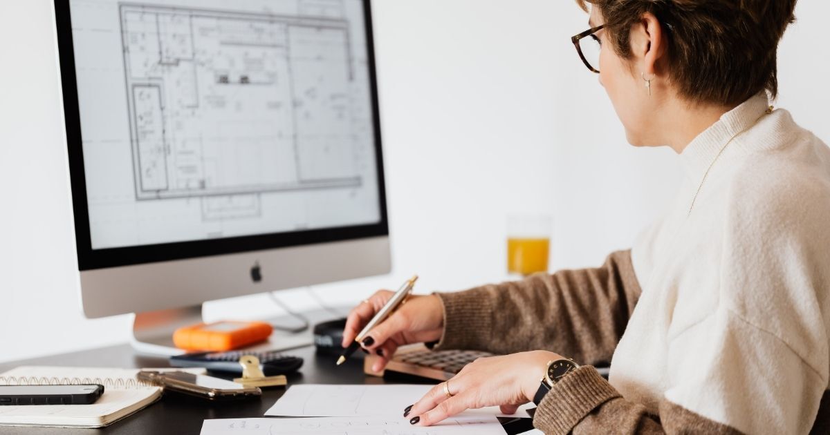 Architect bekijkt een bouwkundige plattegrond op een computerscherm en maakt notities aan haar bureau.