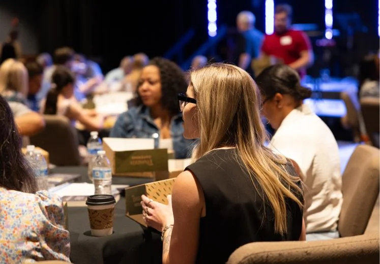 Conference attendees seated at tables with coffee and water bottles
