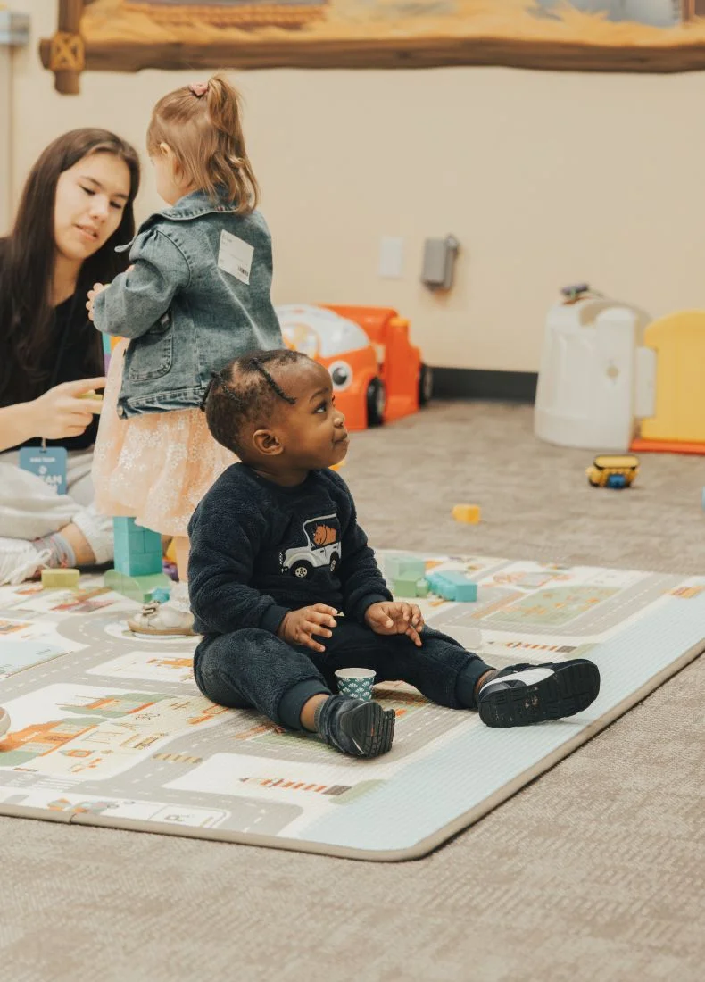 Two children play together on a colorful playmat with toys and caregiver