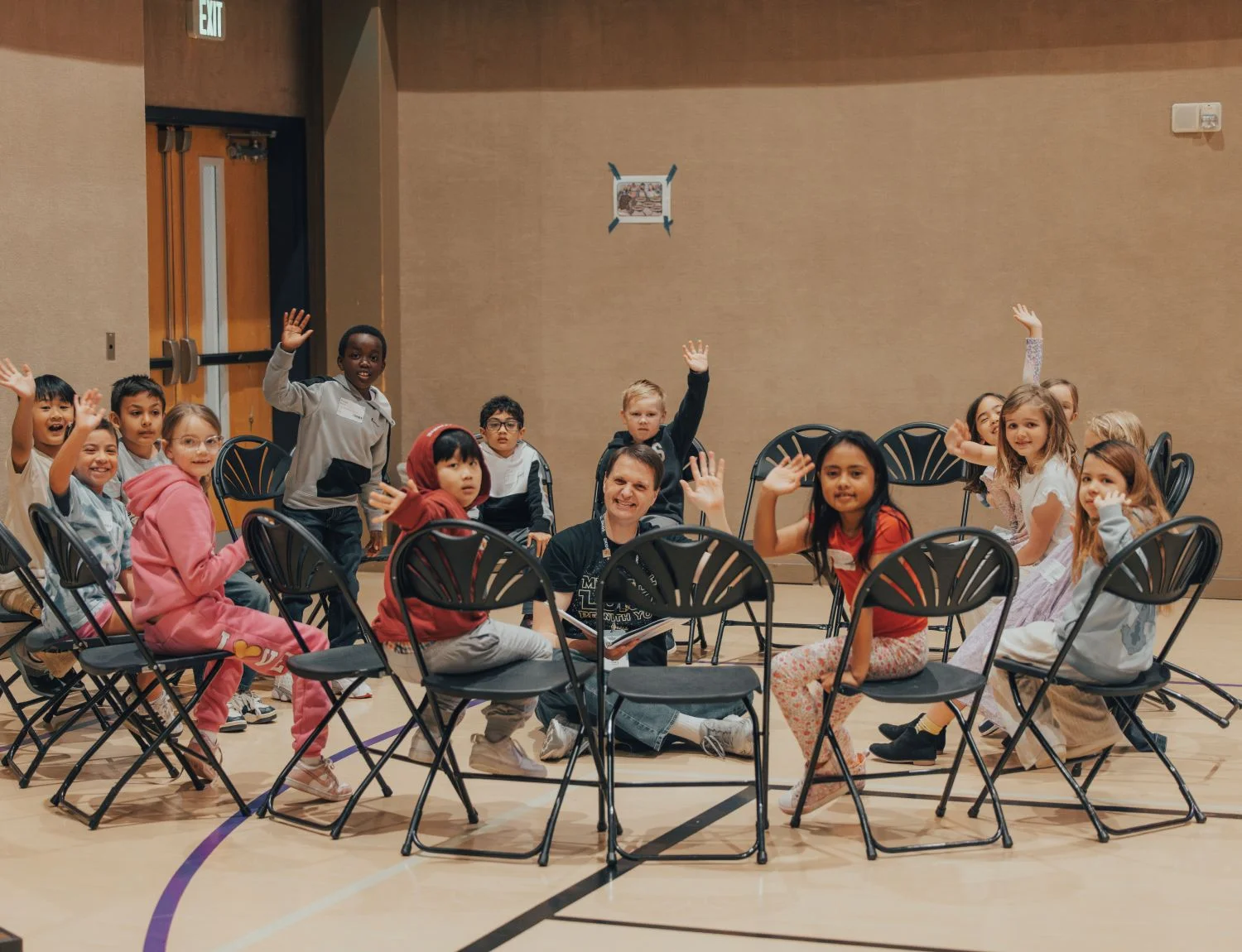 Diverse group of children raising hands while sitting in folding chairs