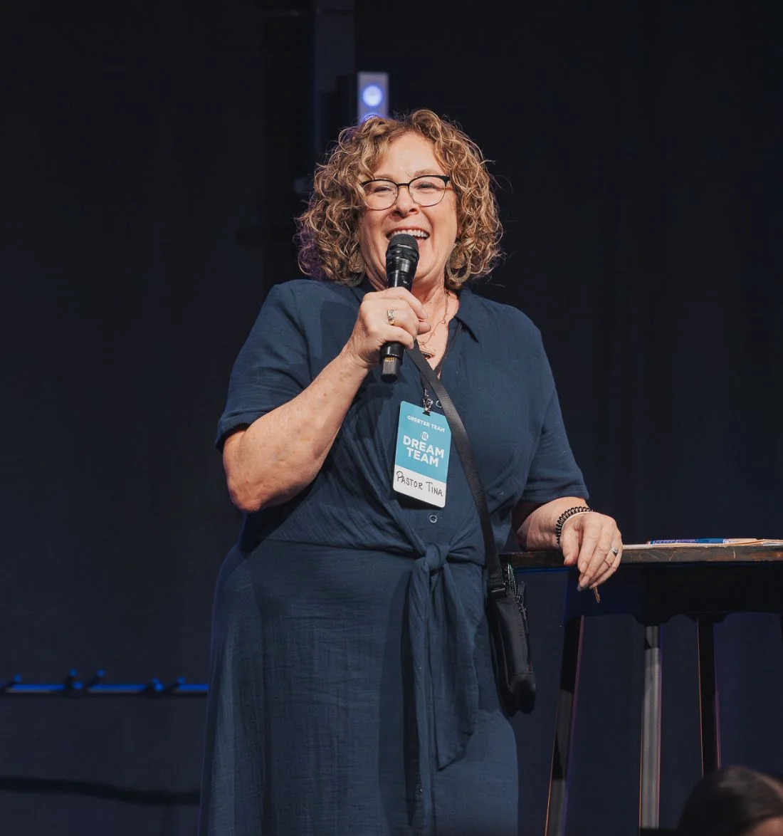 Curly-haired speaker laughing on stage with microphone, wearing Dream Team badge