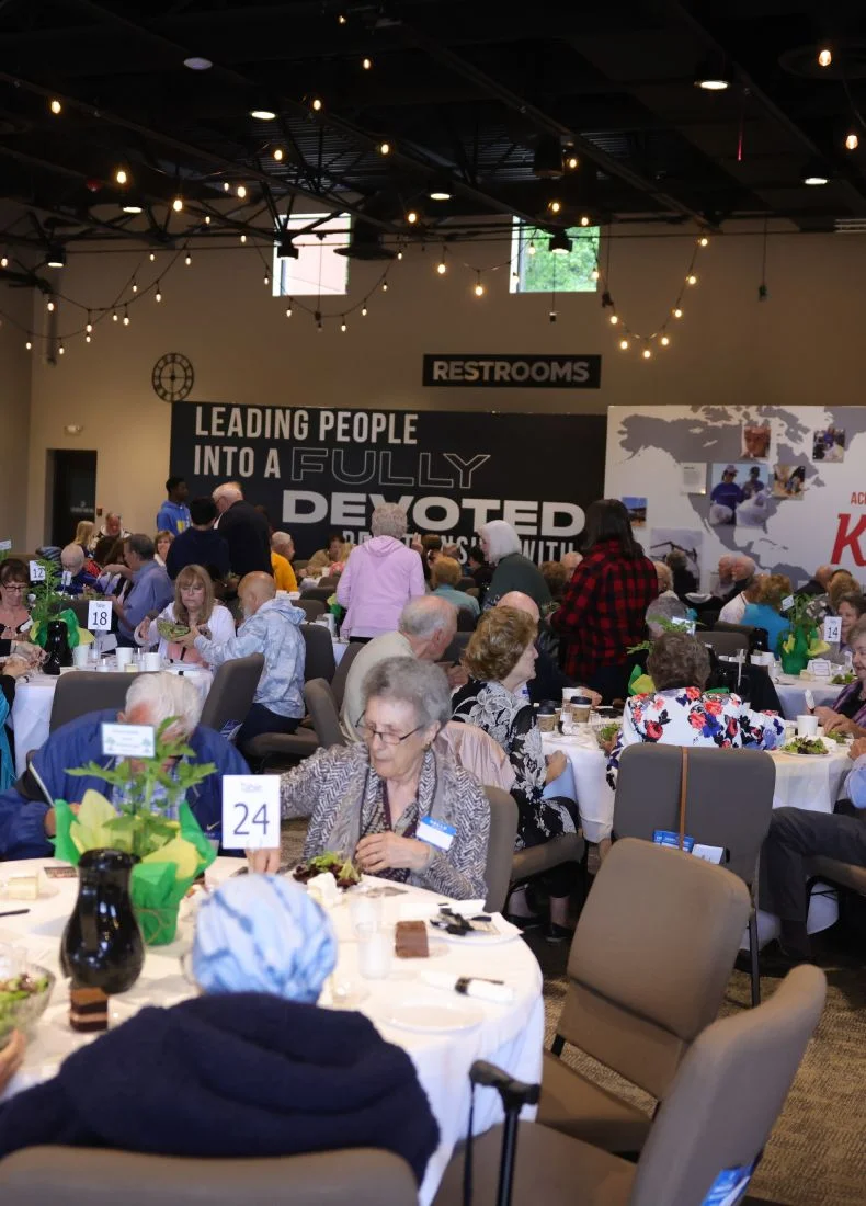 Conference room with string lights, tables, and attendees at a church event