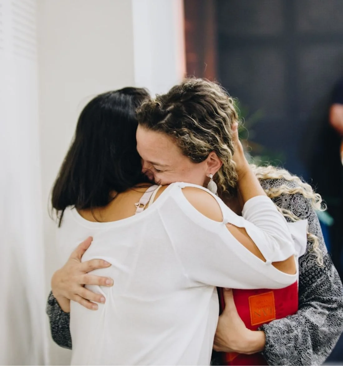 Two women hugging emotionally, one wearing white and holding a red book