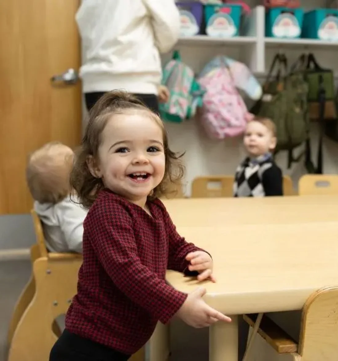 Smiling toddler in red sweater standing at table in daycare classroom