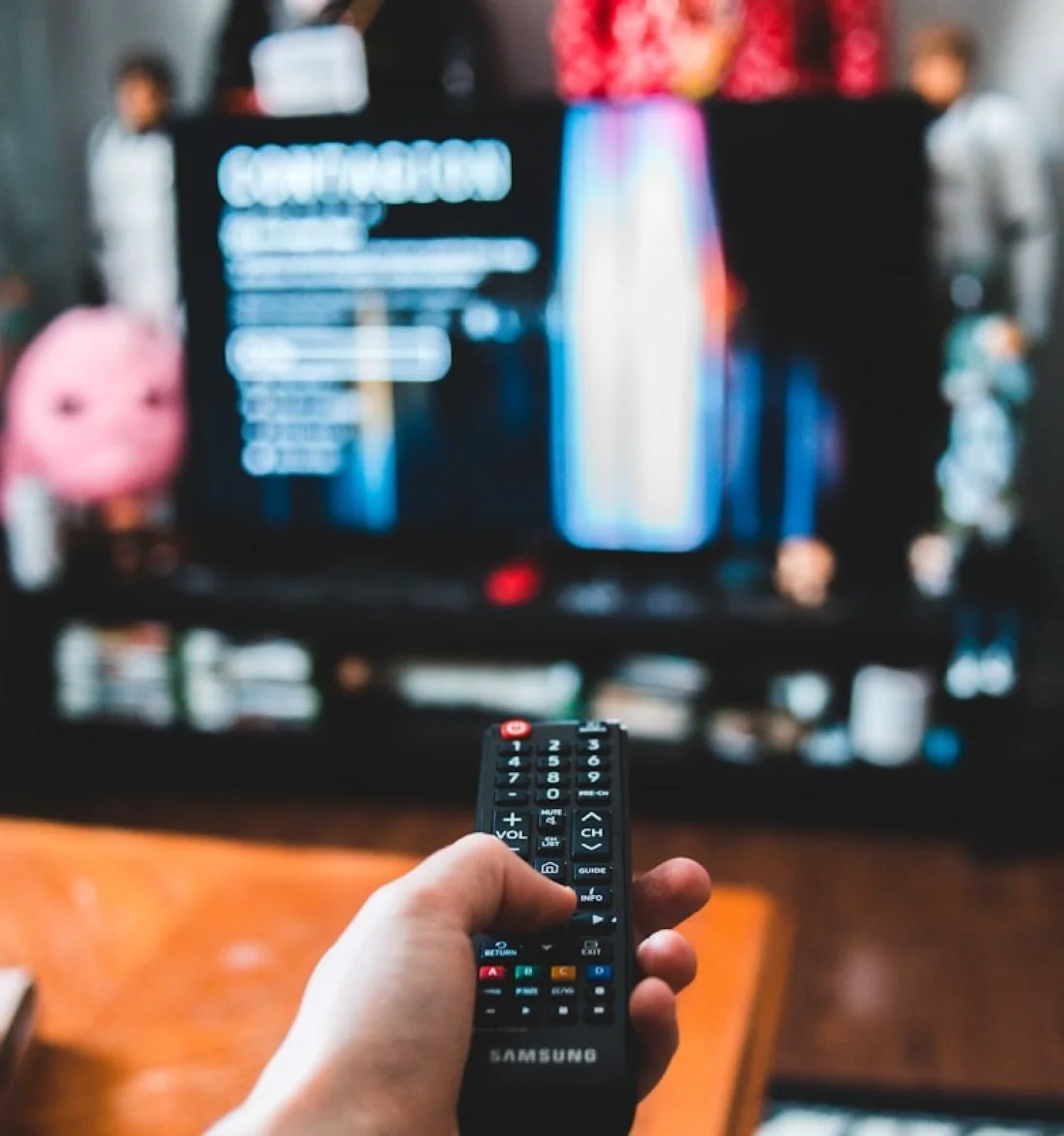 Hand holding Samsung remote control in front of blurry television screen