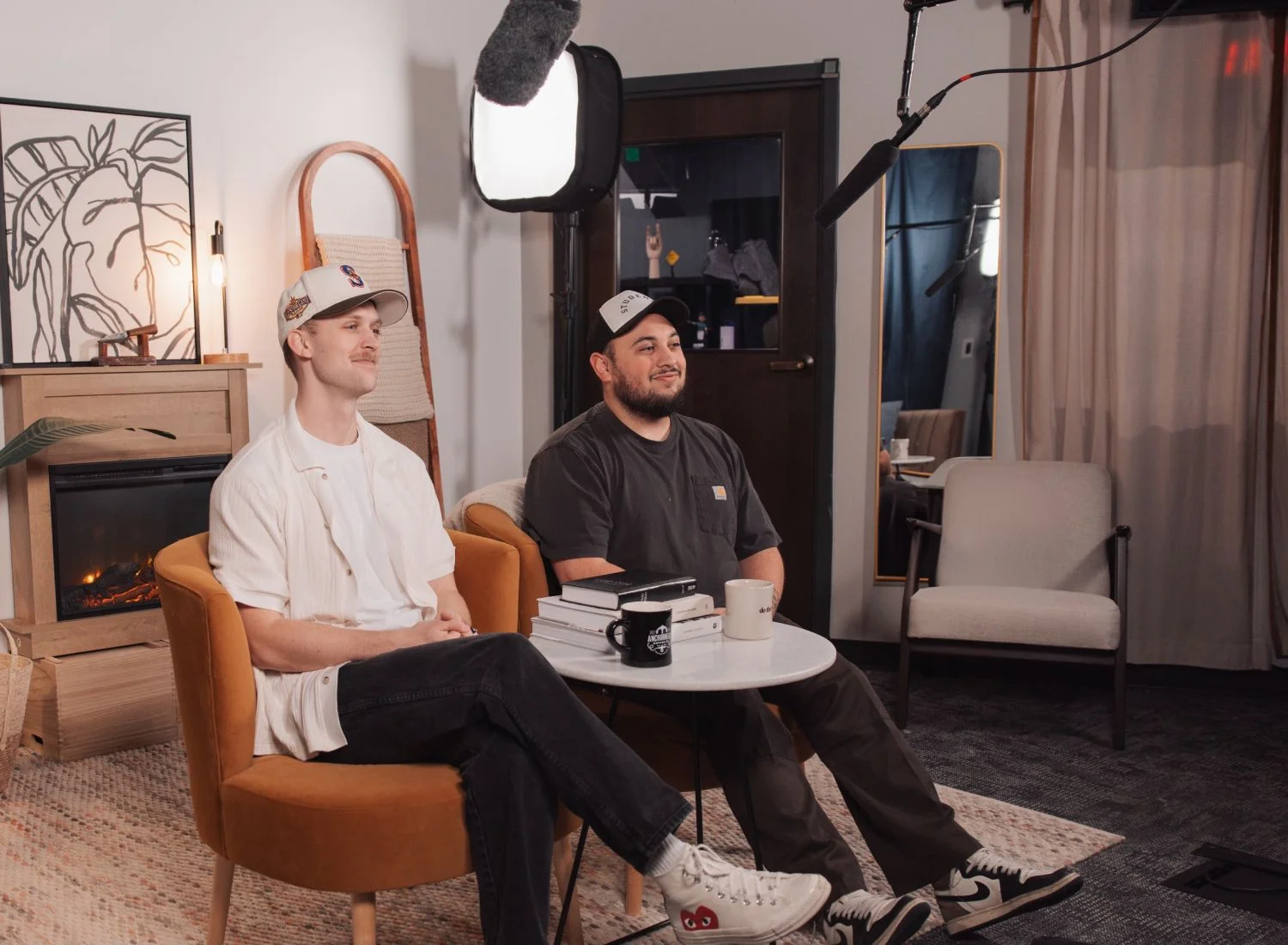 Two men in caps sitting in a cozy studio setting with coffee and books