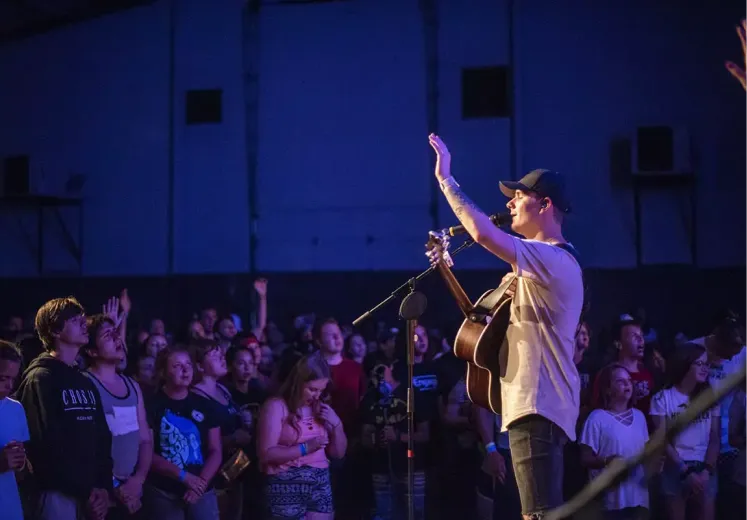 Musician performing on stage with raised hand, acoustic guitar, enthusiastic crowd