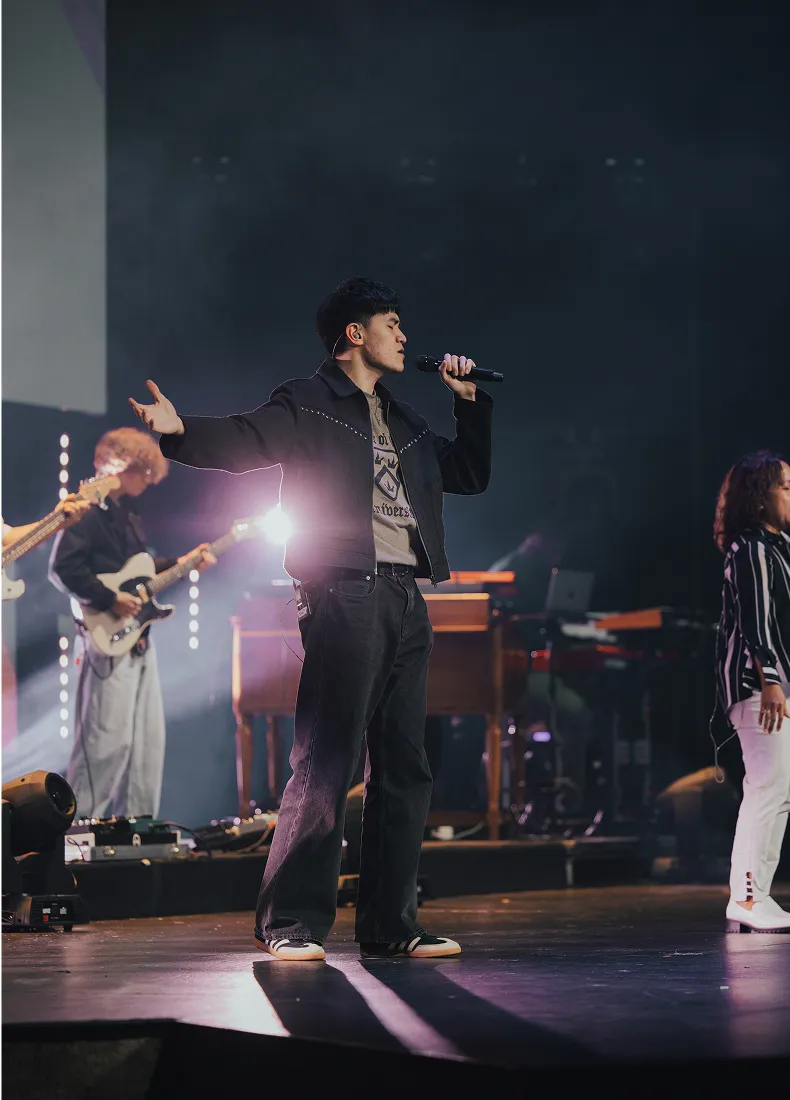 Singer performs on stage with band, microphone in hand, dramatic lighting