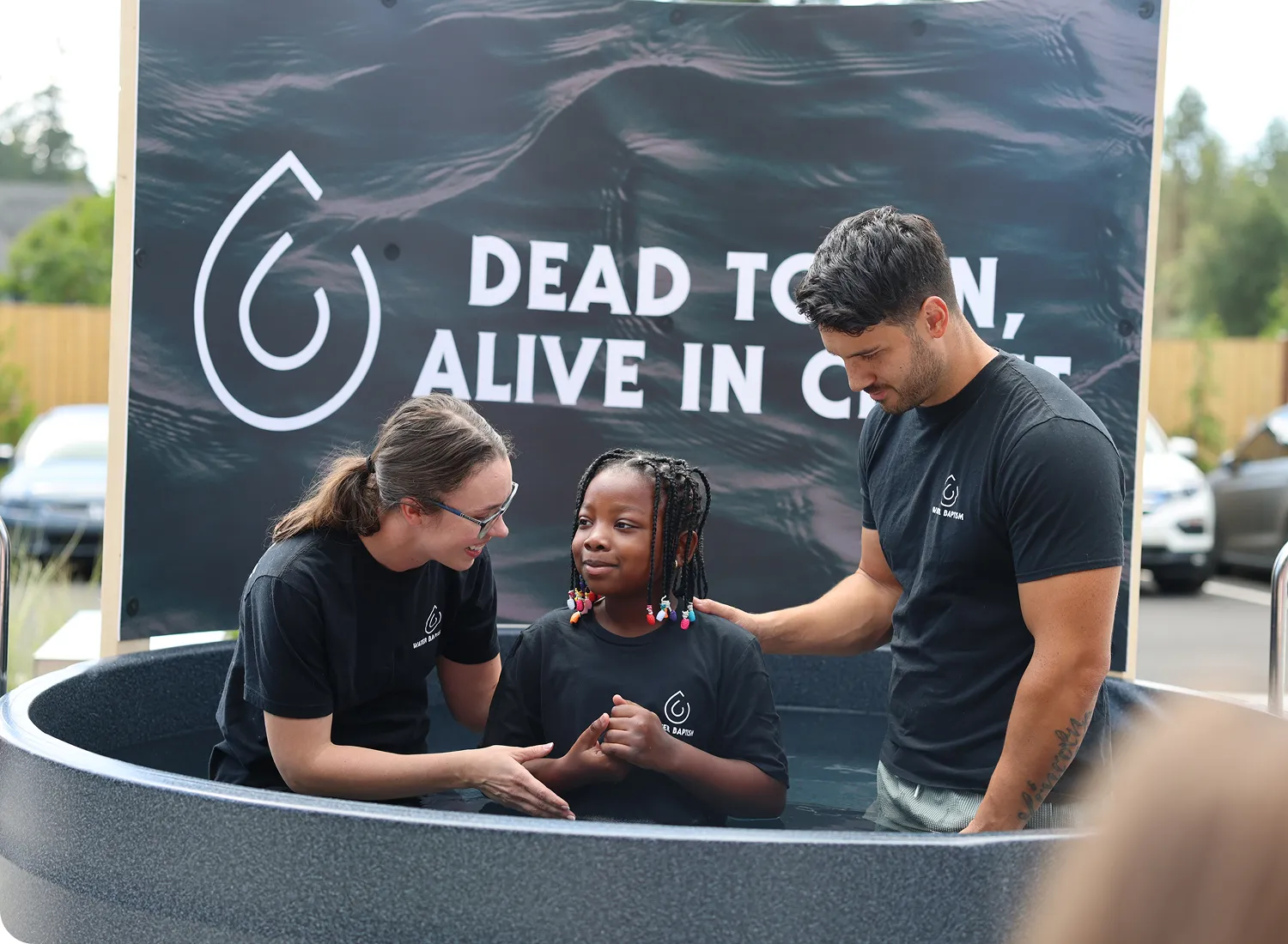Three people in black shirts at baptism event with "Dead to Sin" banner