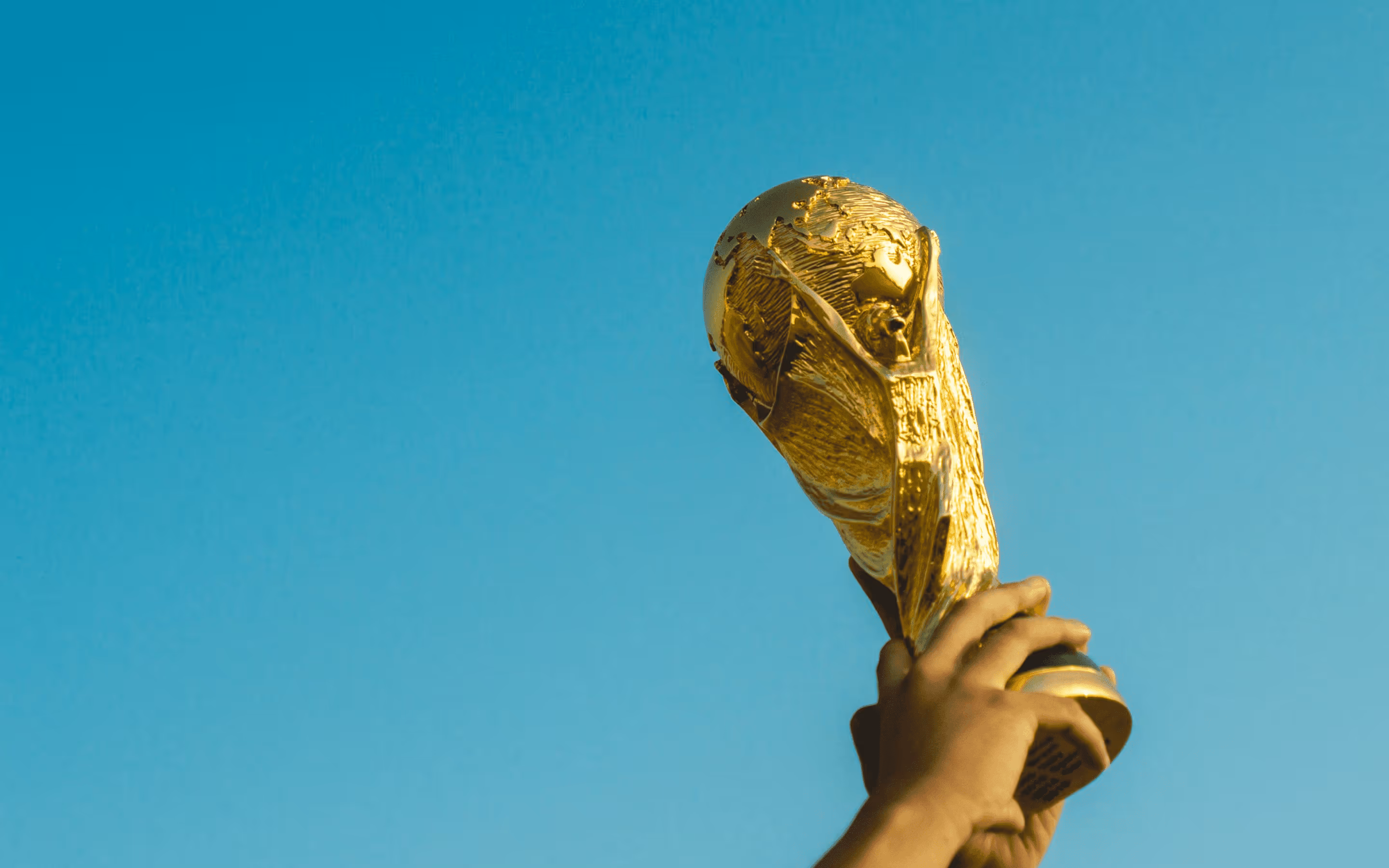 Two hands holding up the FIFA World Cup trophy against a clear blue sky.