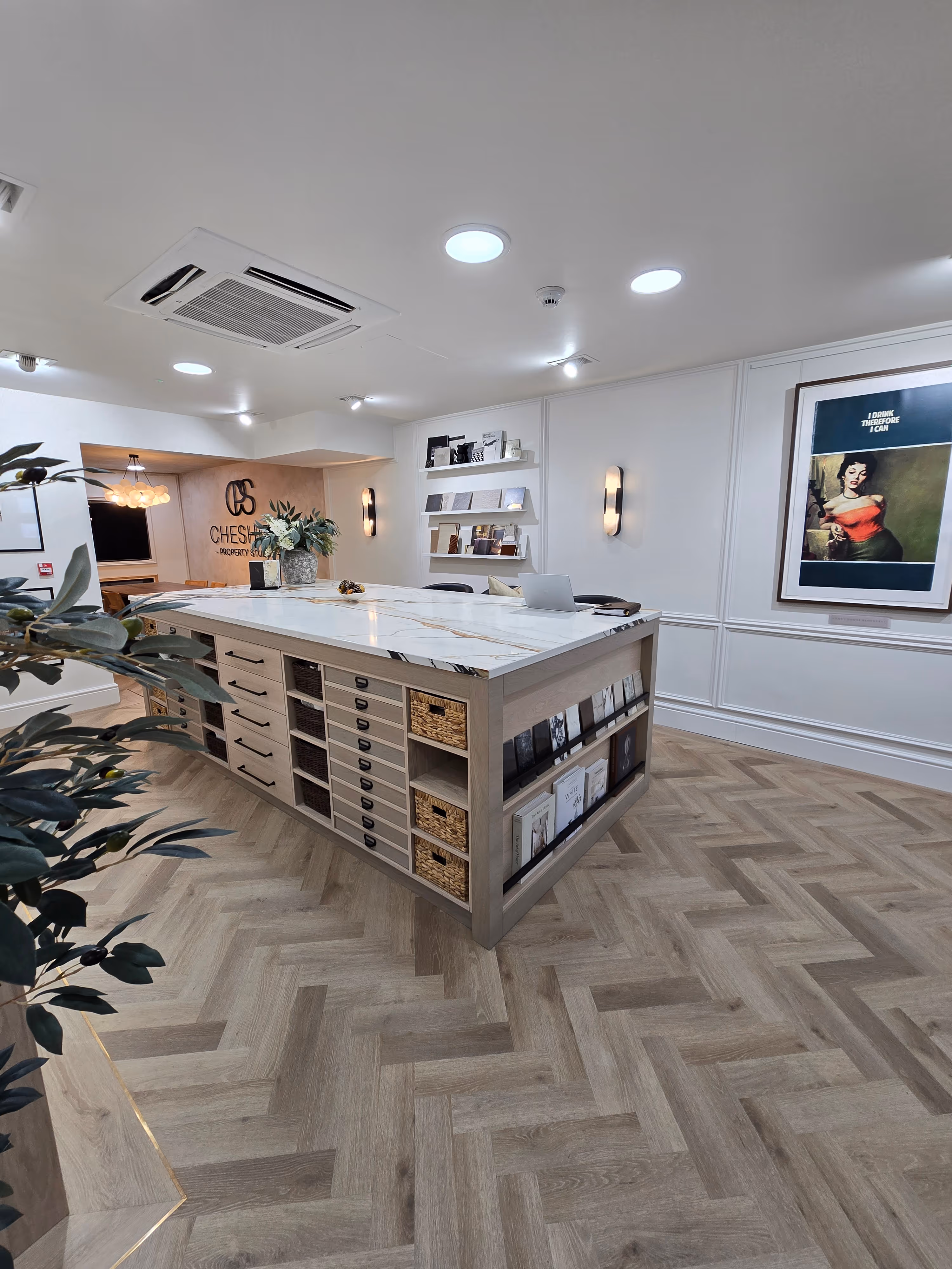 Modern office interior with a large marble-topped worktable, wooden herringbone floor, white walls, and shelving displaying books and samples.