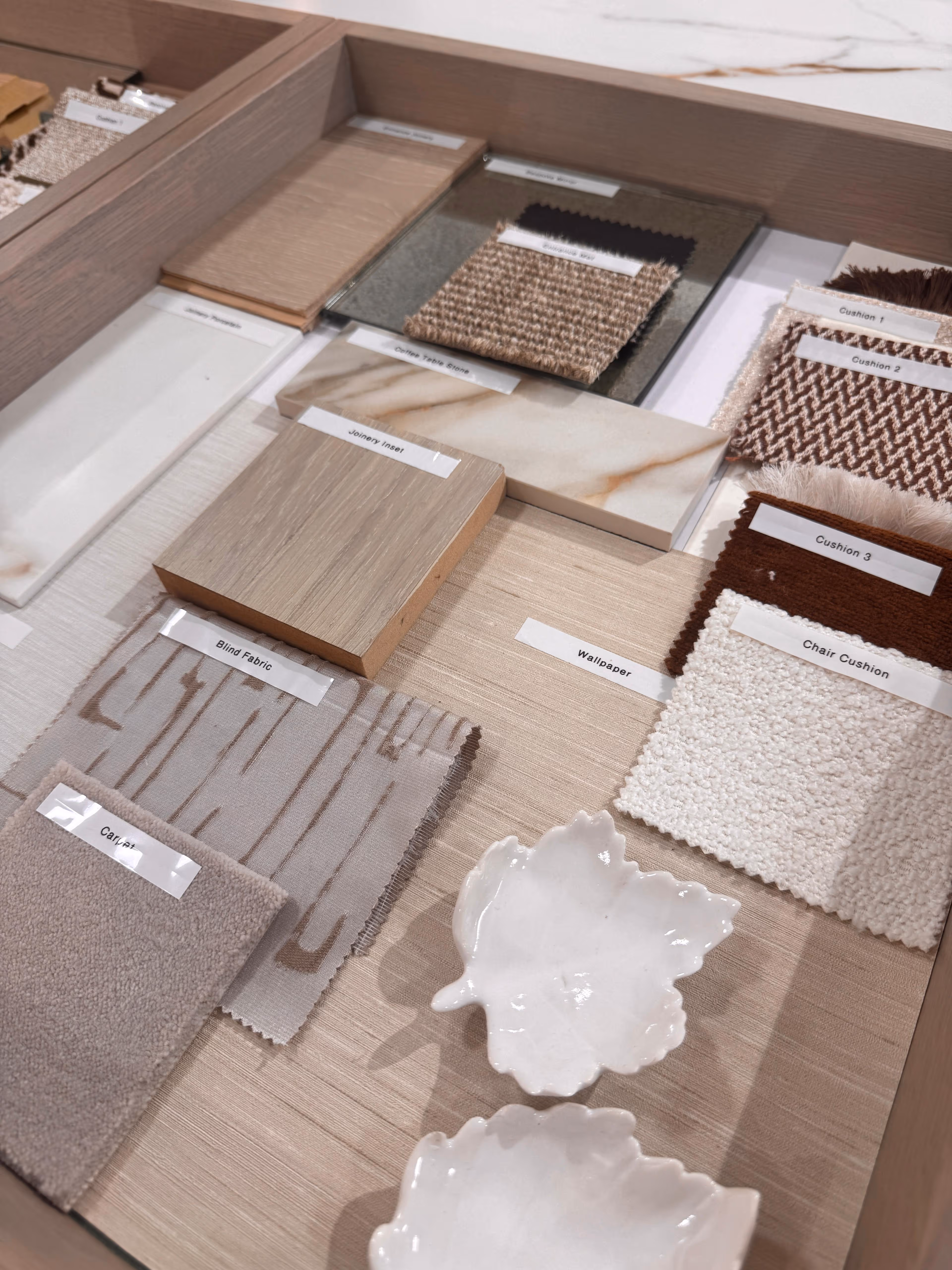 Sample board with fabric swatches, carpet, wallpaper, marble and wood samples, and two white ceramic leaf-shaped dishes arranged on a light wood surface.