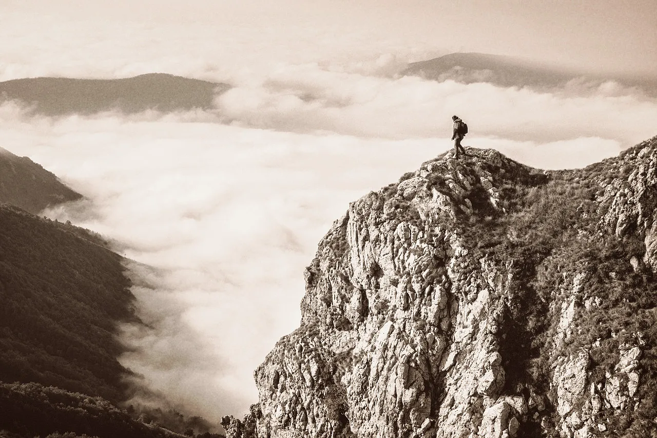 Hiker standing on a rocky mountain peak overlooking a valley filled with clouds and distant hills.