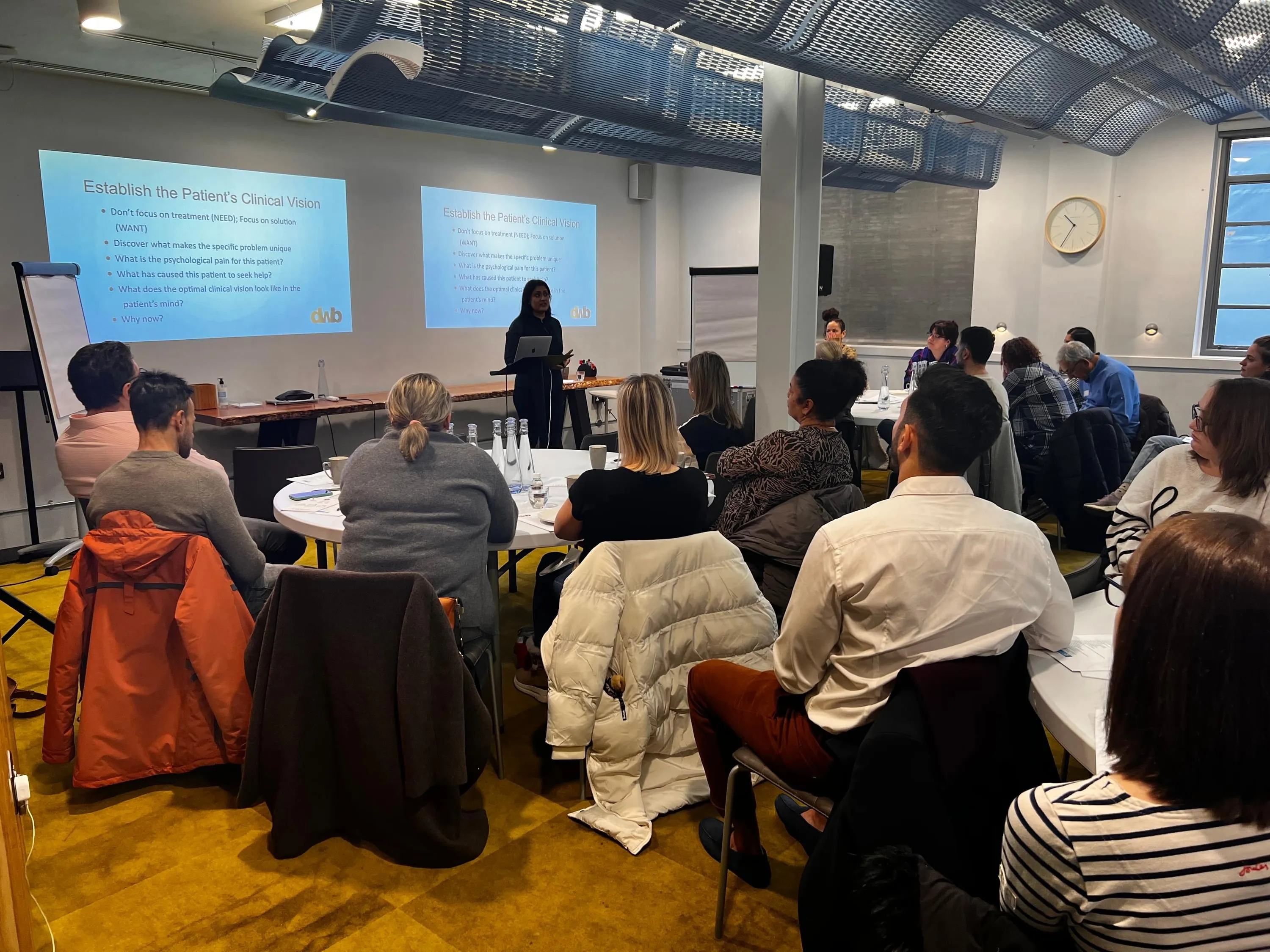 Speaker presenting slides to a seated room of dental professionals at a training workshop