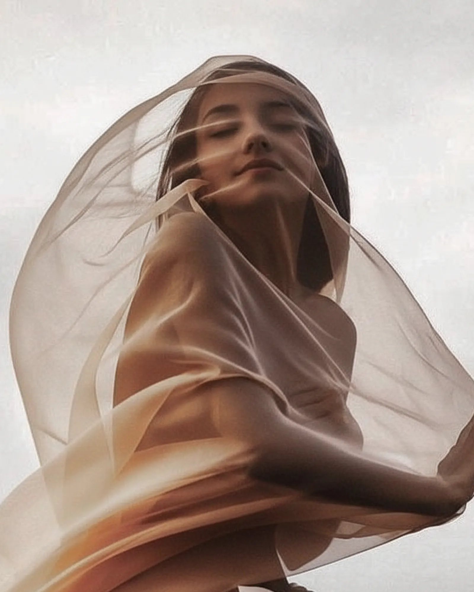 A woman with closed eyes wrapped in flowing translucent beige fabric against a pale sky.