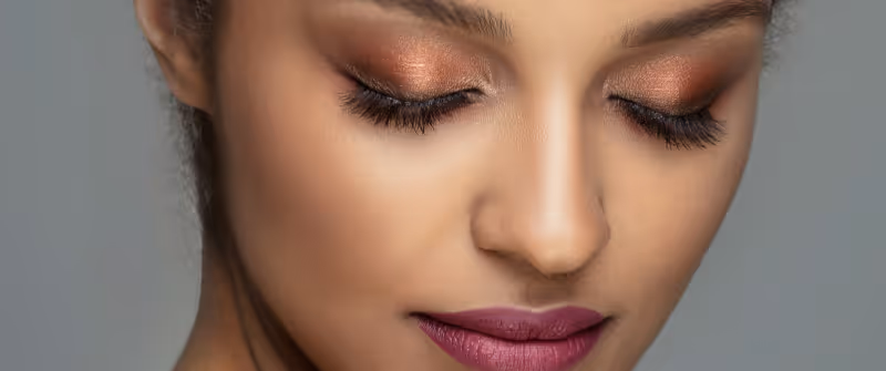 Close-up of a woman with copper-colored eyeshadow, long eyelashes, and pinkish-mauve lipstick on her closed eyes and lips.