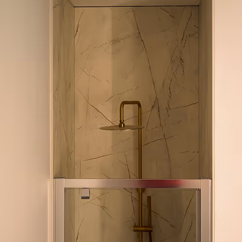 Modern shower with brass fixtures and beige marble walls behind a glass door.