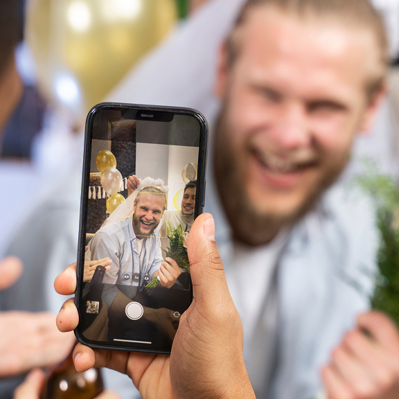 Person holding a smartphone taking a photo of a laughing man wearing a veil and holding flowers at a celebration with gold balloons in the background.