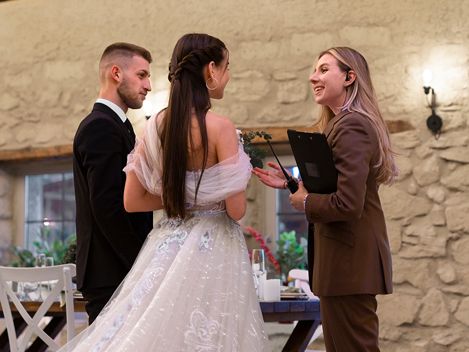 Wedding couple talking with a woman in a brown suit holding a clipboard inside a rustic venue.