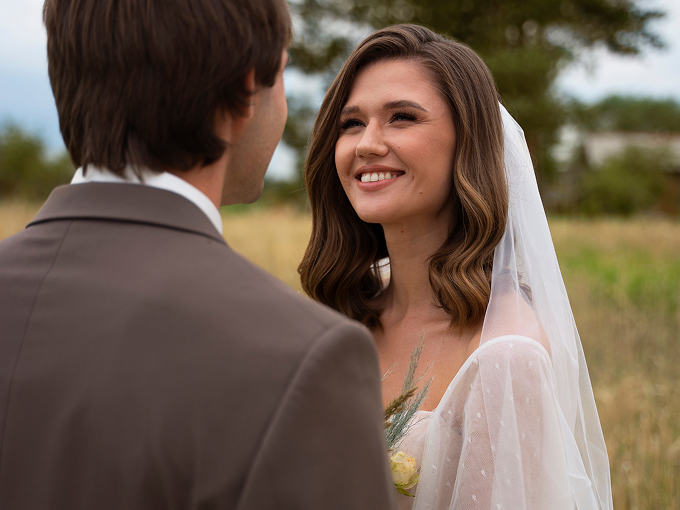 Bride smiling at groom outdoors, wearing a white veil and dress.