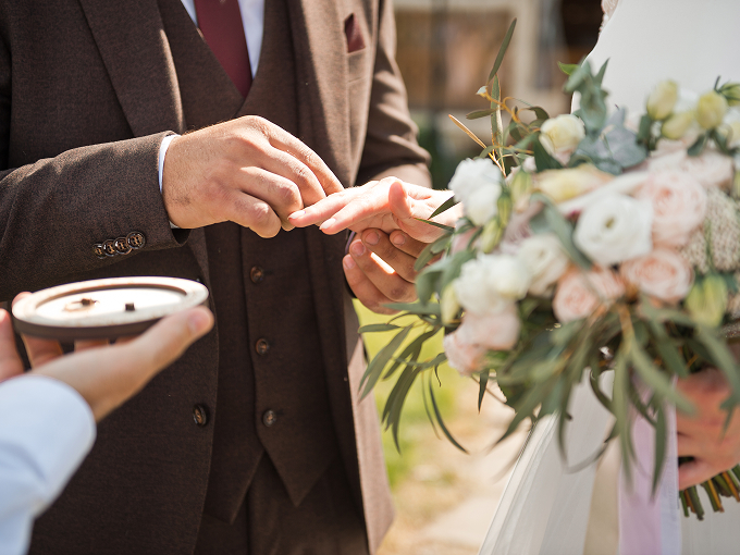 Groom placing a wedding ring on bride's finger as she holds a bouquet of white and blush flowers.