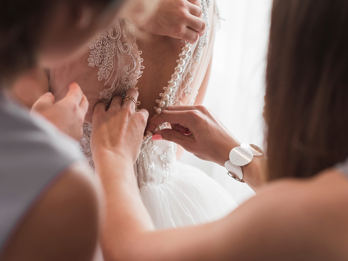 Close-up of two women fastening buttons on the back of a lace wedding dress.