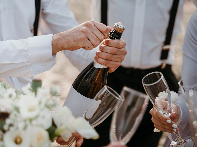 Person in white shirt opening a champagne bottle with others holding champagne flutes and white flowers in the foreground.
