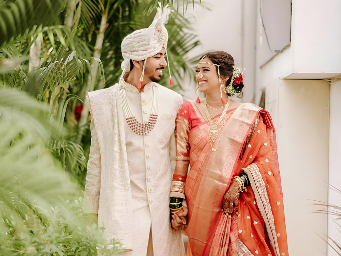 Indian bride and groom holding hands and smiling at each other amidst greenery.