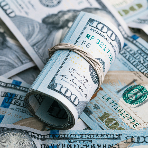 Rolled bundle of US 100 dollar bills secured with a rubber band resting on scattered hundred-dollar notes.