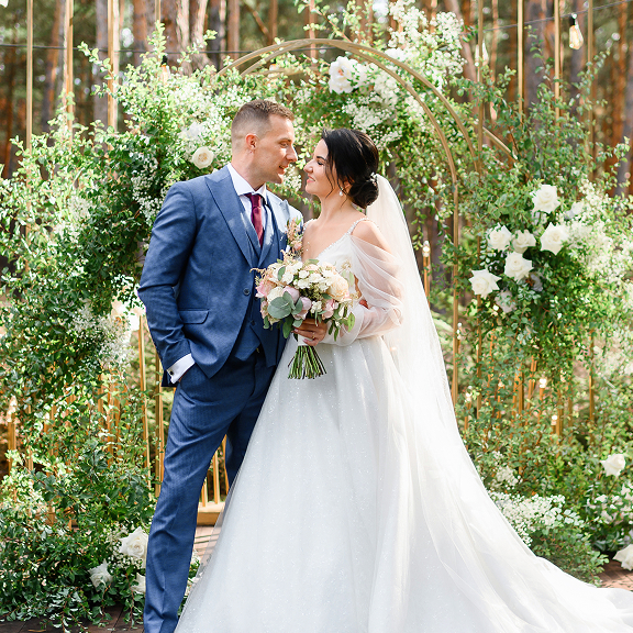 Bride in white gown holding bouquet and groom in blue suit standing closely in front of floral arch.