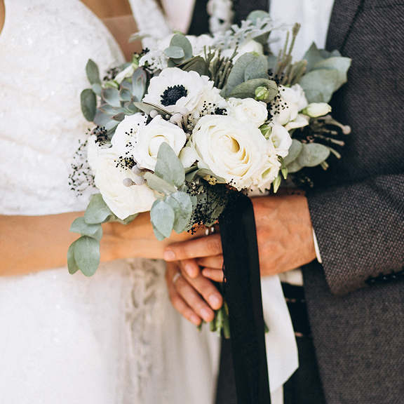 Bride and groom holding a bouquet of white flowers with green leaves and a black ribbon.