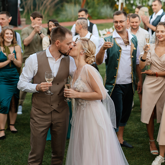 Newlywed couple sharing a kiss while holding champagne glasses during an outdoor celebration with guests applauding in the background.