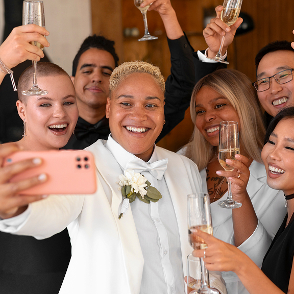 Group of six smiling friends dressed formally, holding champagne glasses and taking a selfie together.