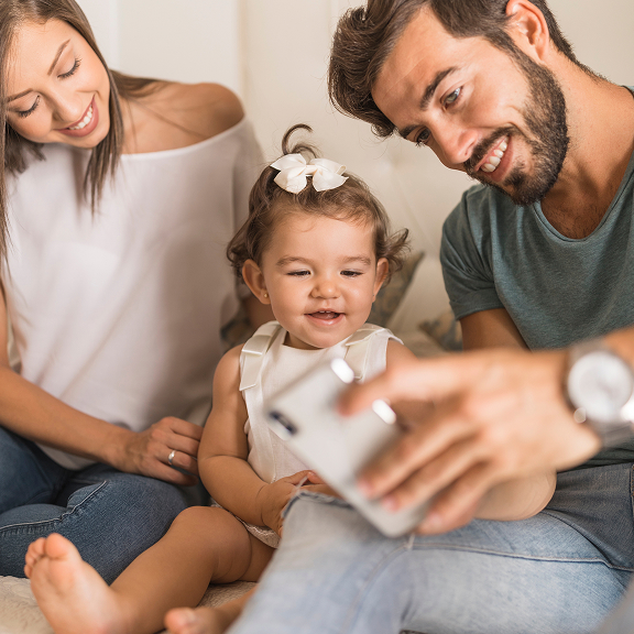 Smiling parents taking a selfie with their young daughter who is wearing a white dress and bow in her hair.