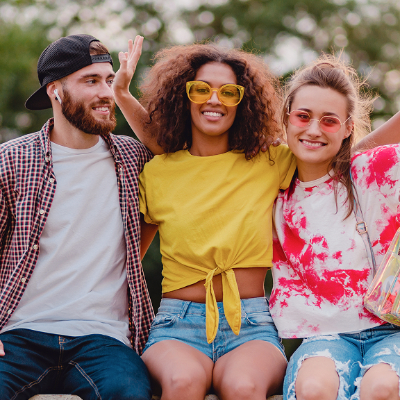 Three friends outdoors smiling, with a man wearing a backward cap and earbuds, a woman in a yellow top and glasses, and another woman in a red and white tie-dye shirt and sunglasses.